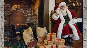 Santa sitting on a chair next to a fireplace and lots of presents wrapped in brown paper and tied up with red ribbon on the floor