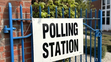 'polling station' sign on a blue fence next to a brick wall and a tree